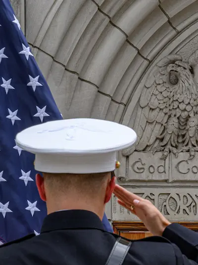 A Marine in a dress uniform and white cover salutes an American flag, with a stone carving of an eagle and the words “God Country Notre Dame” in the background.